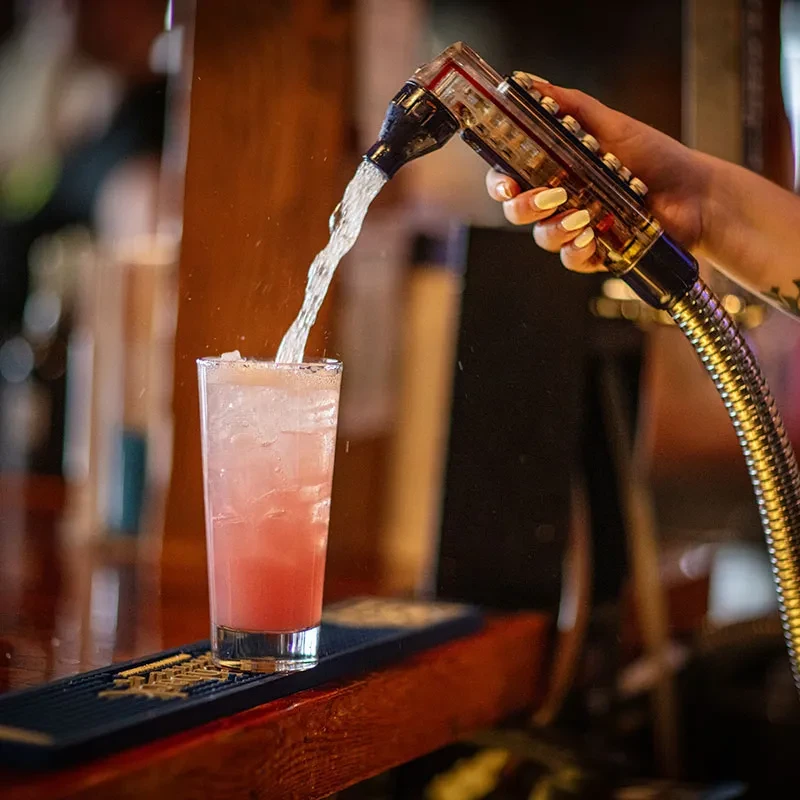 Hand dispensing clear liquid from soda gun into pink drink, ice.