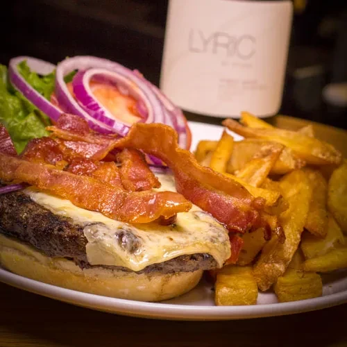 Close-up bacon cheeseburger and wedge fries with a LYRIC bottle.