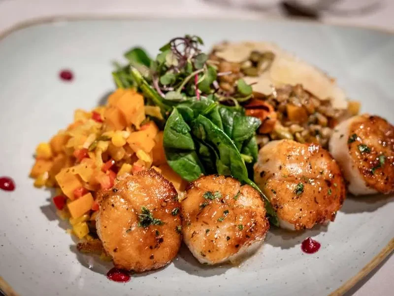 Plated dish: four seared scallops, colorful vegetables, greens, and parmesan risotto.