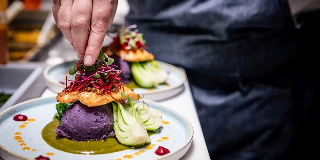 Chef's hand adds microgreens to plated fish, purple purée, bok choy.