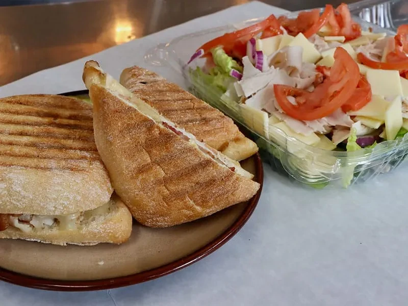 Two paninis on a plate next to a to-go salad container.