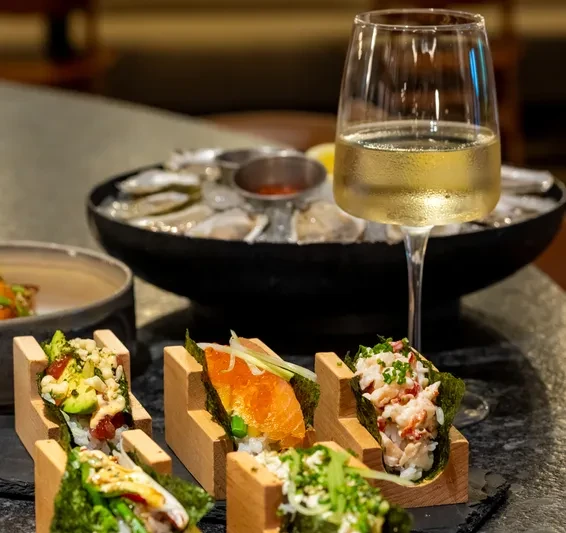 Four hand rolls, raw oysters on ice, and wine on a dark table.