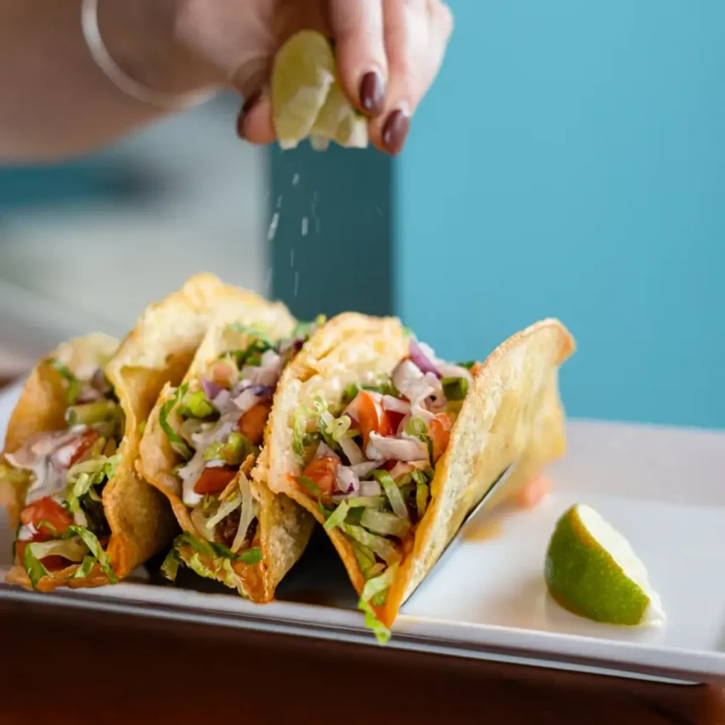 Hand squeezing lime over three crispy tacos with fresh toppings.