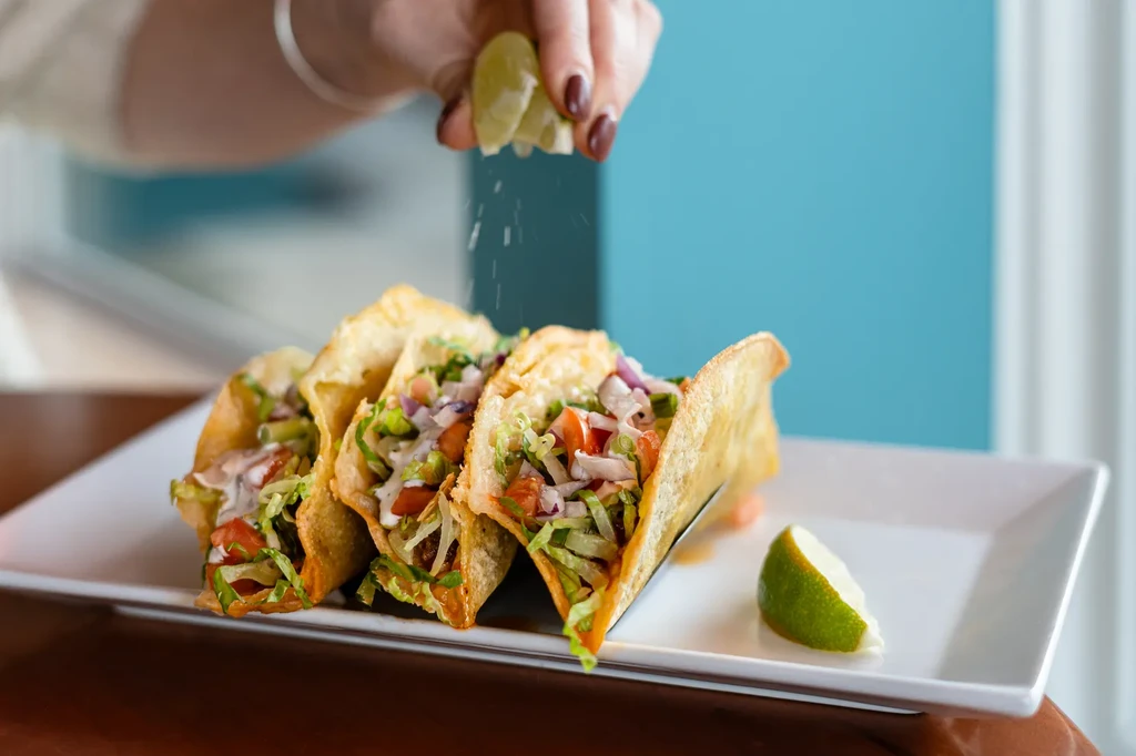 A hand squeezes lime over three crispy, filled hard-shell tacos.