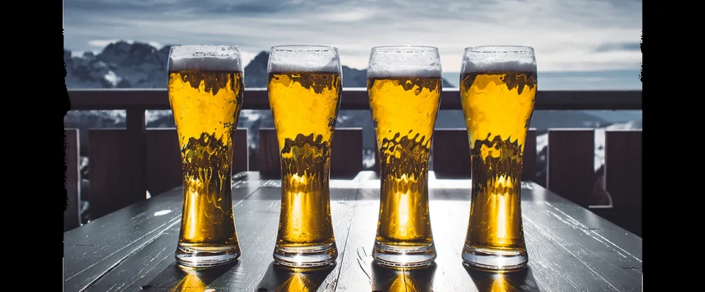 Four pilsner beers on a railing overlooking snow-capped mountains.