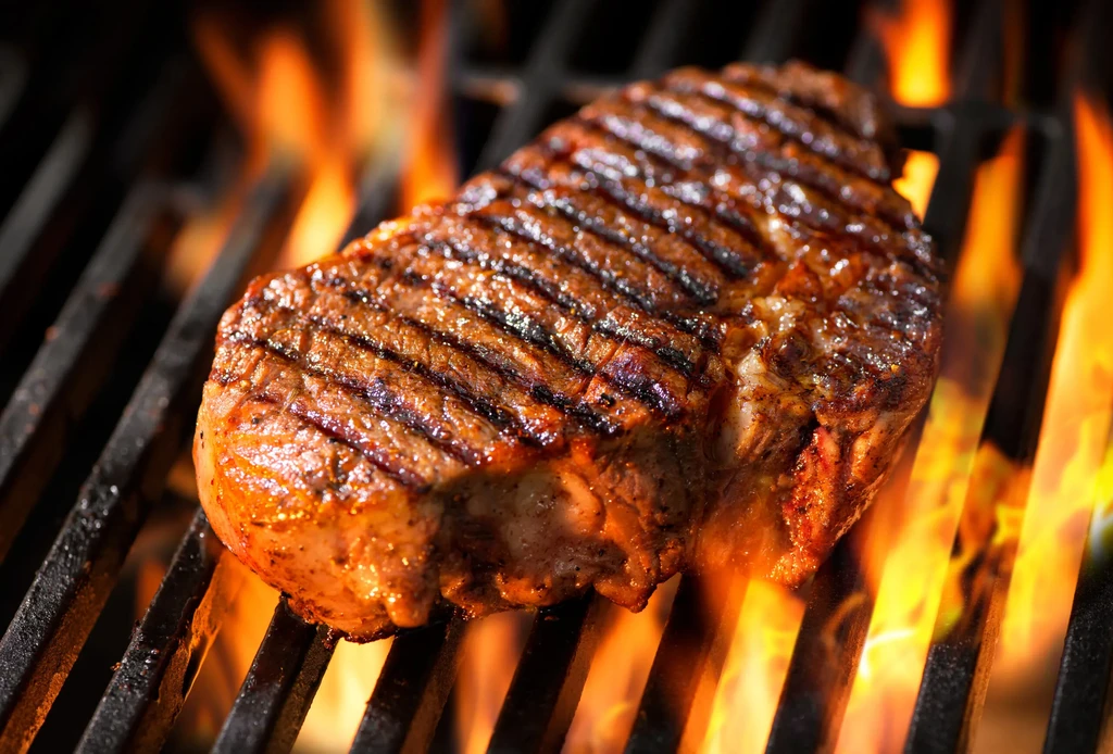 Thick cut steak grilling over vibrant open flames with grill marks.