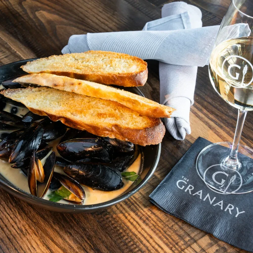 Mussels in creamy broth, baguette, and white wine on a wooden table.