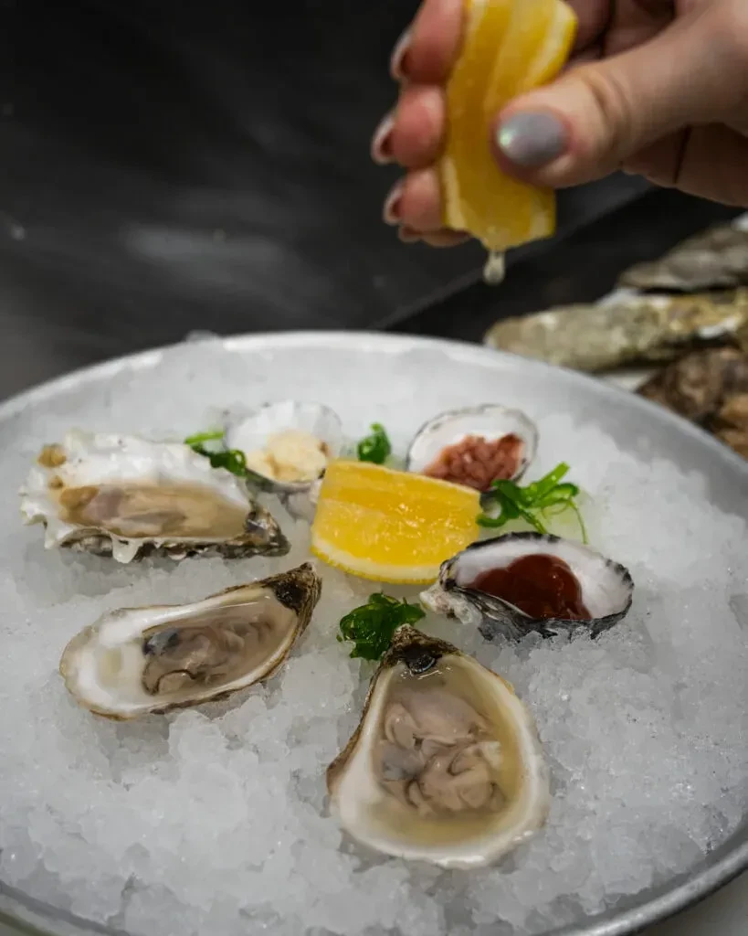 A hand squeezes lemon over fresh oysters served on crushed ice.