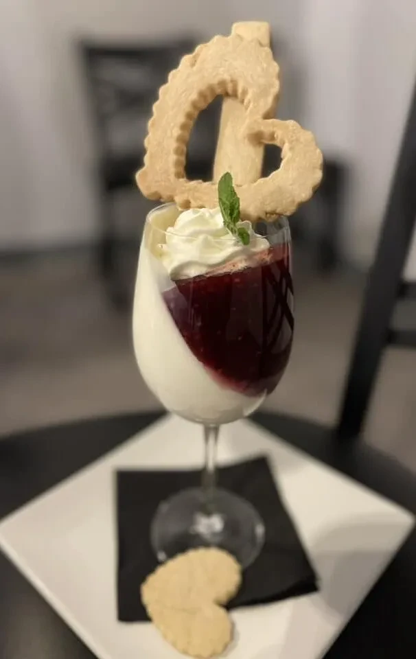 Layered dessert in wine glass with fruit, whipped cream, mint, heart cookie.