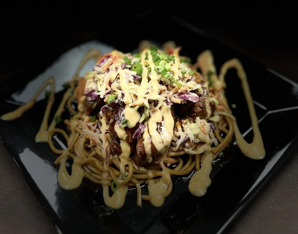 Noodles, dark meat, colorful slaw, and creamy sauce on a black plate.