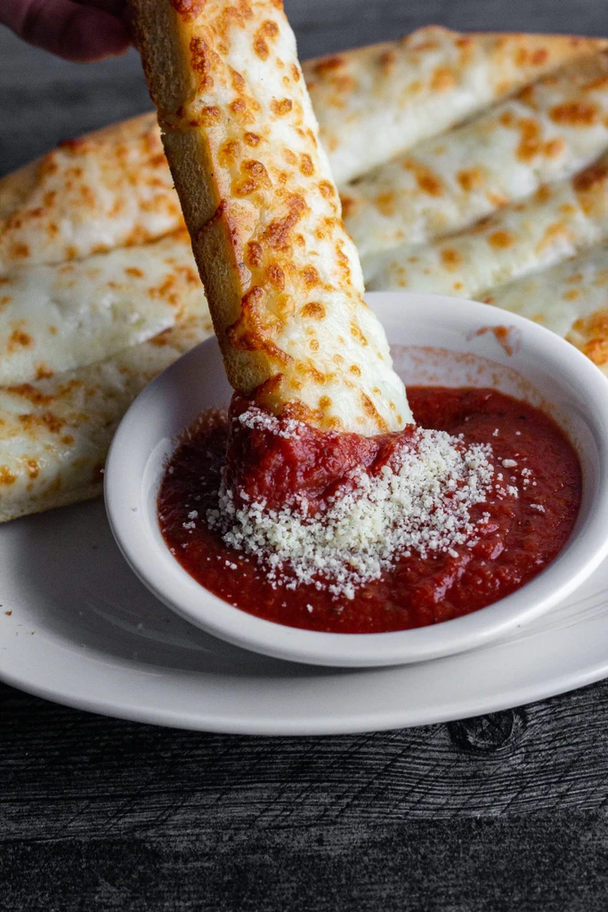 Golden cheesy breadstick dipping into marinara sauce topped with cheese.