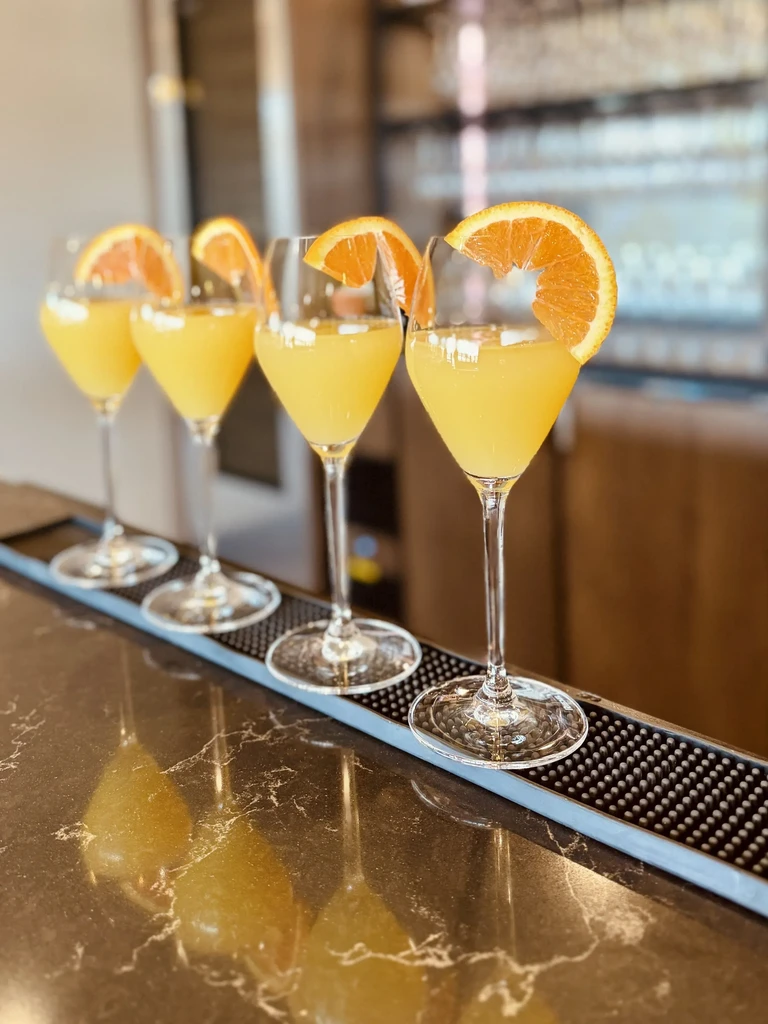 Four mimosas with orange slices in flutes on a reflective bar.