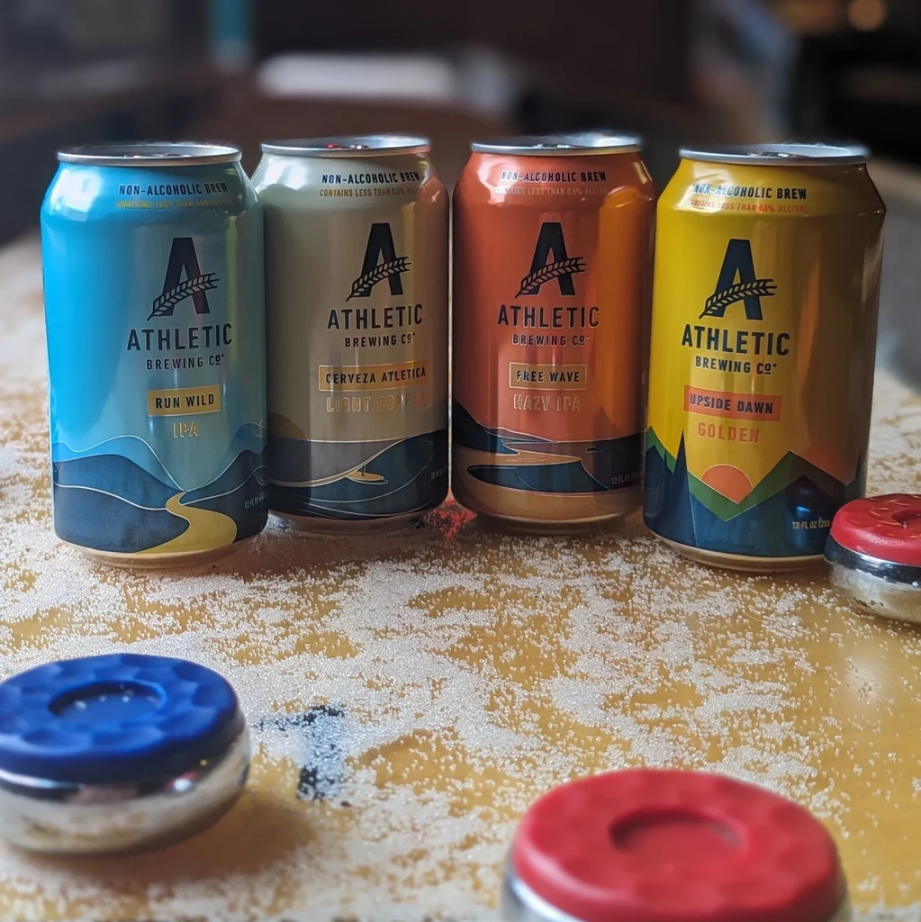 Four non-alcoholic Athletic Brewing cans and shuffleboard pucks on a table.