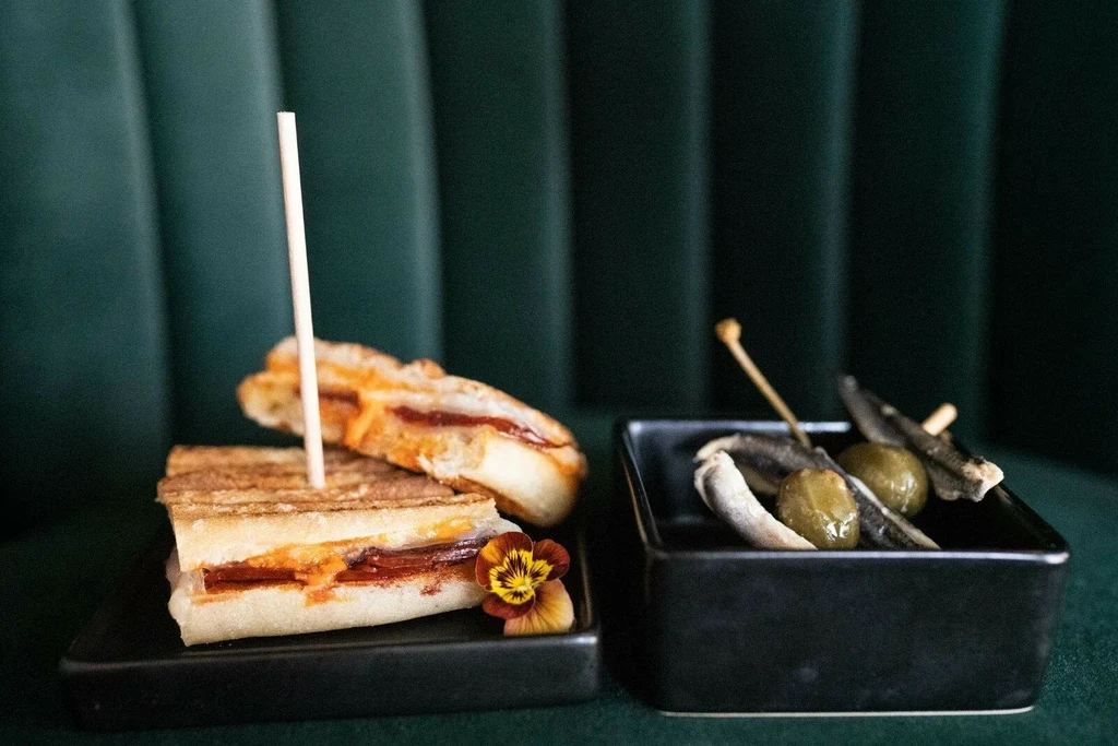 Pressed sandwich, olives, and pickled fish on two black dishes.