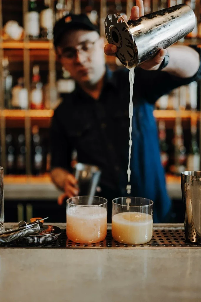 Bartender pours cocktail from shaker into one of two foamy drinks.