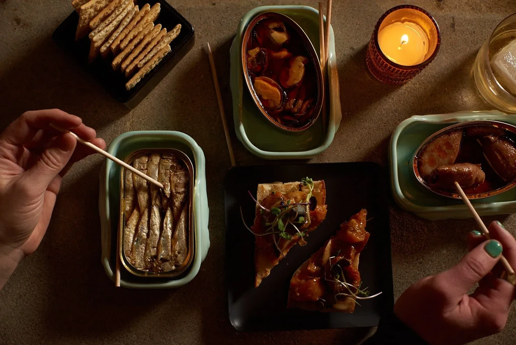 Hands hold skewers over various seafood appetizers, crackers, drinks, and candle.