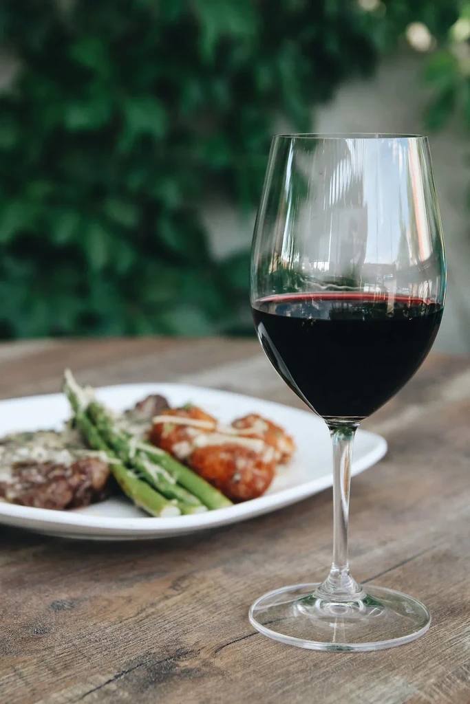 Red wine glass on wood table with blurred meal and green foliage.