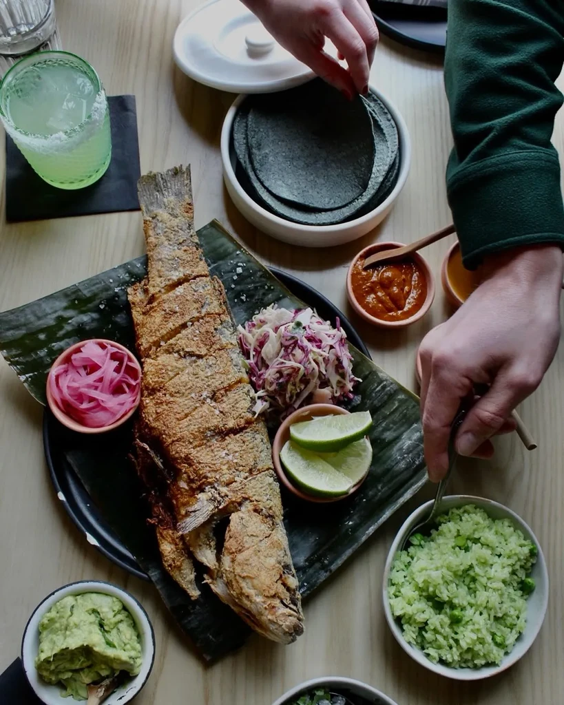 Overhead photo of fried fish, green rice, blue tortillas, and drinks.