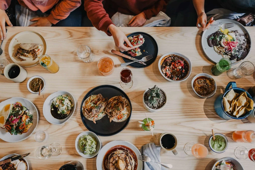 Overhead: Diverse food and drinks on a table with hands reaching.