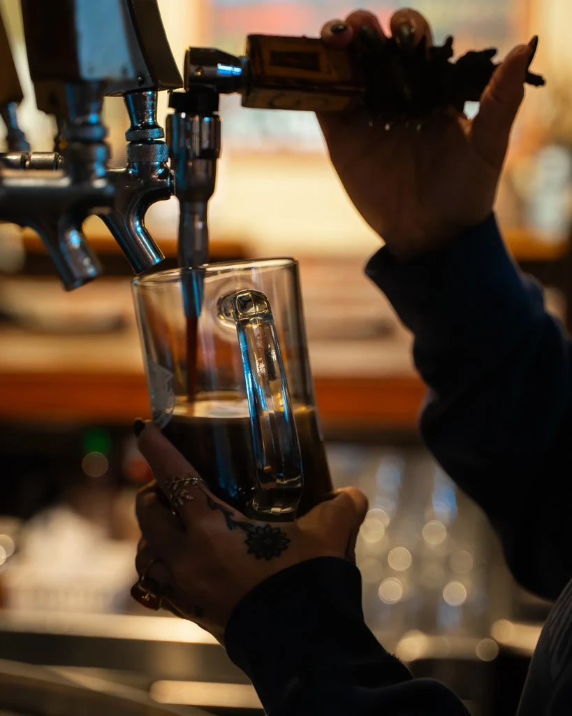Hands with rings and tattoo pour dark beer from a tap.