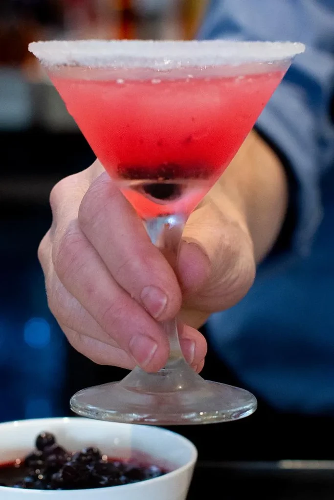 Reddish-pink cocktail in martini glass with sugared rim, held by hand.