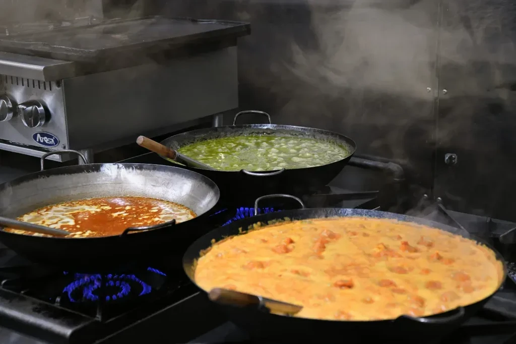 Three large woks bubbling with diverse dishes on a commercial stove.