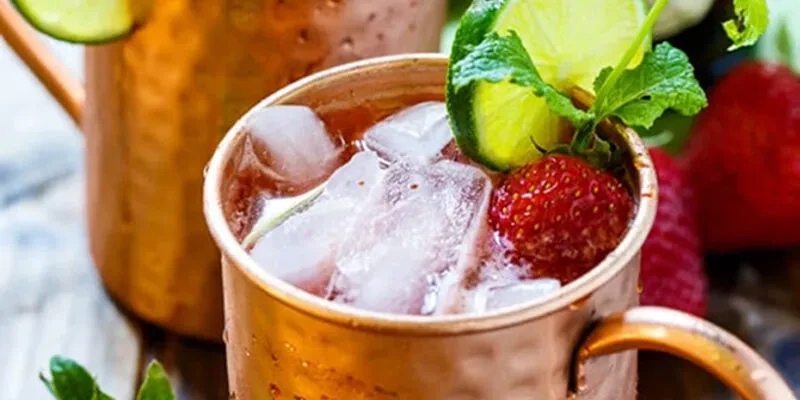 Reddish-brown iced cocktail in copper mug, with lime, mint, strawberry.