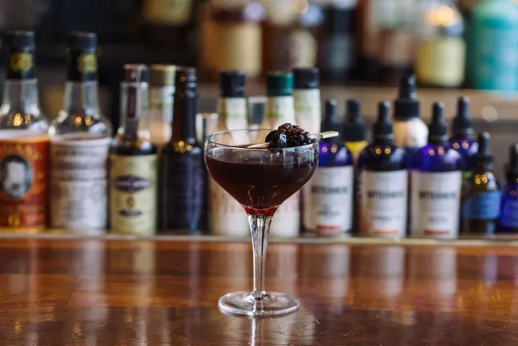 Reddish-purple cocktail in coupe glass with blackberries on a bar.