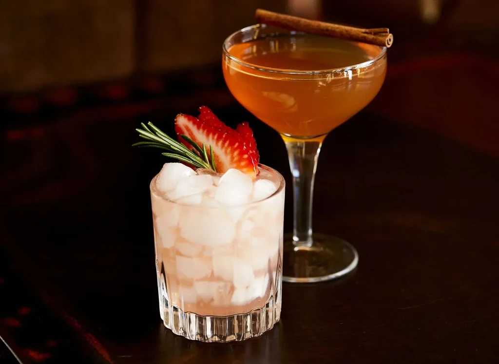 Two garnished cocktails: pink with strawberry, amber with cinnamon stick.