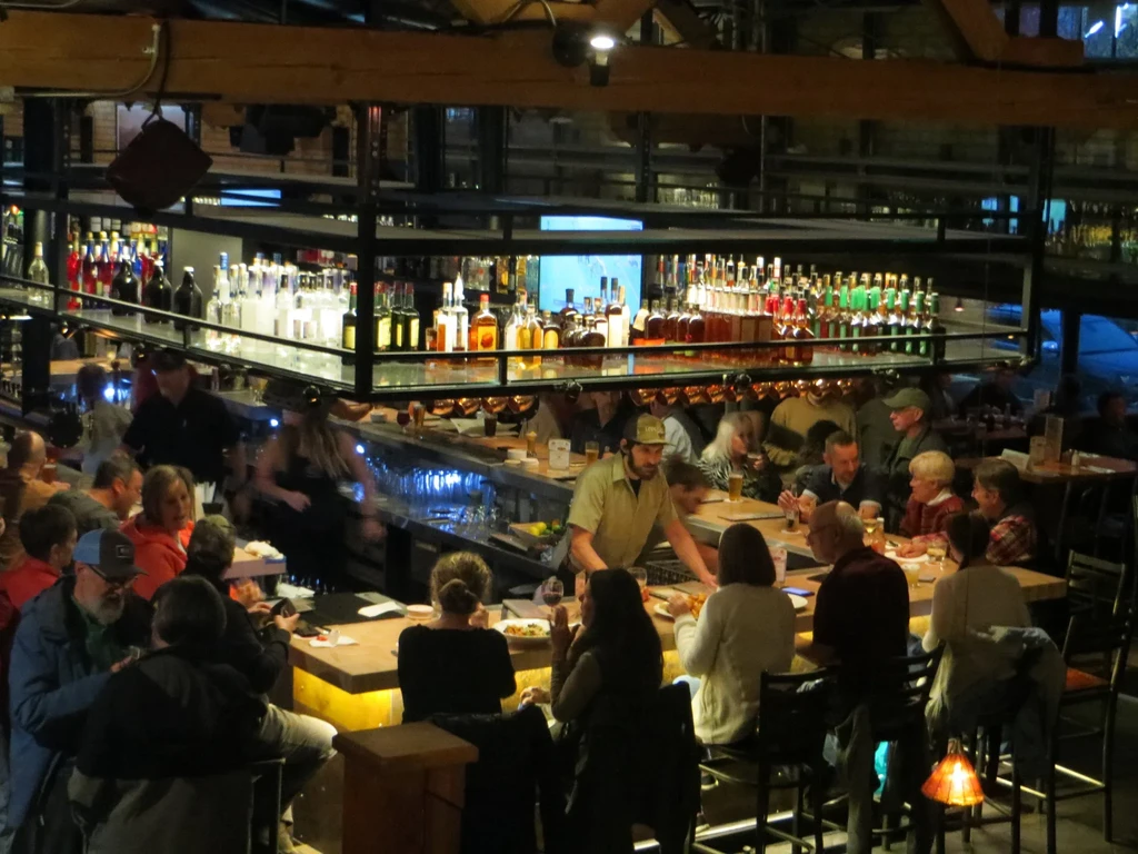 Rustic bar interior, bustling with patrons, staff, and illuminated bottles.