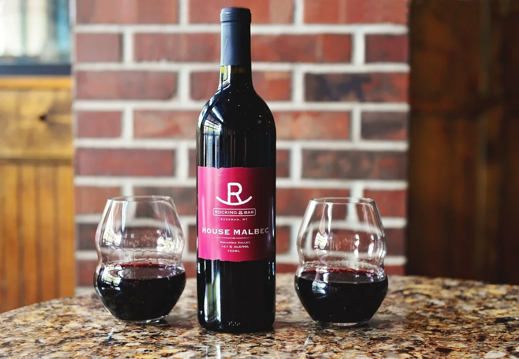 R Rocking J Bar Malbec bottle, two glasses, brick wall background.