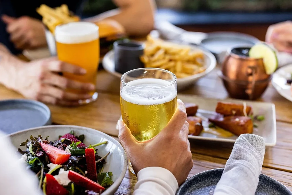 Hand holding a cold beer, salad, and appetizers on a wooden table.