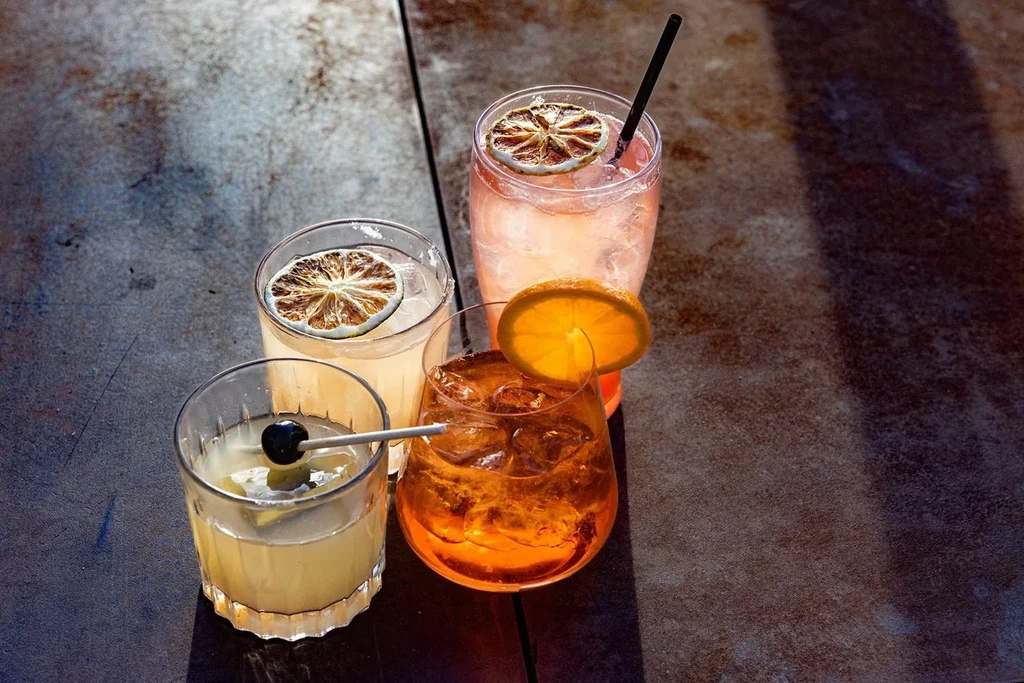 Four colorful, garnished cocktails on a dark surface in bright sunlight.