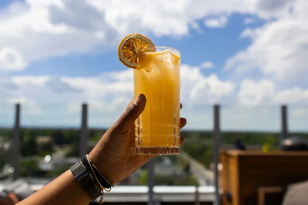 Dark-skinned hand with bracelets holds orange drink, dried slice, under blue sky.