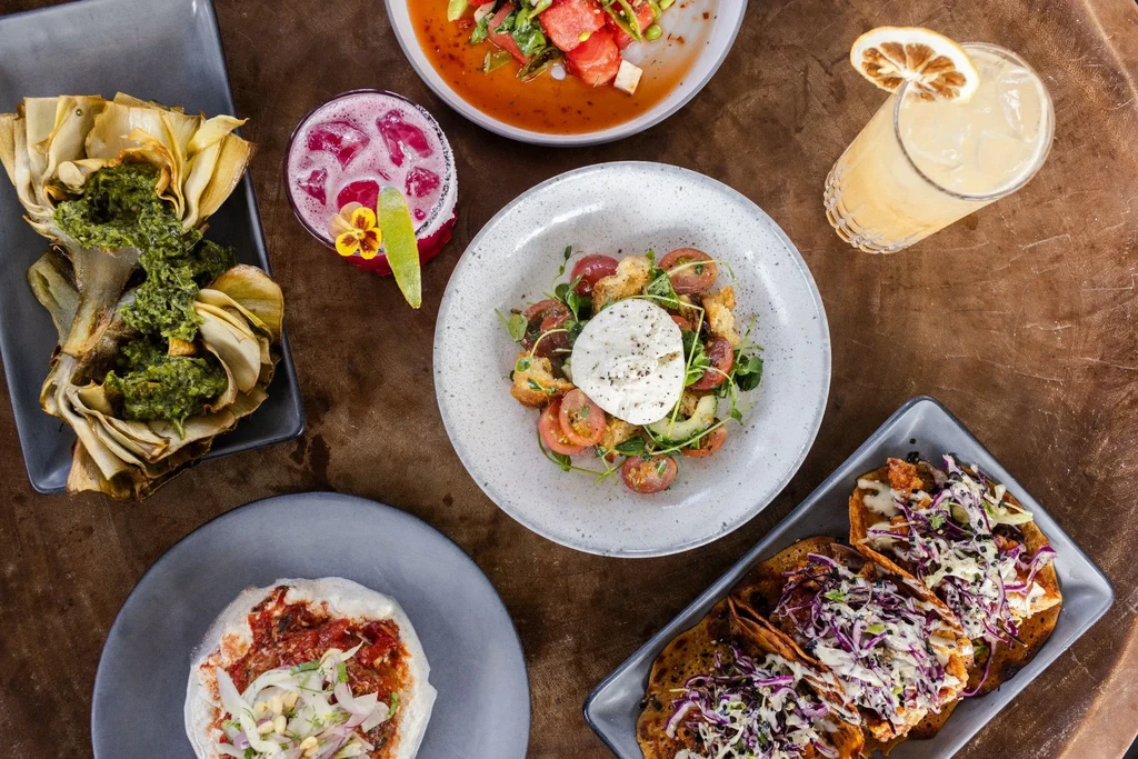 Overhead view of roasted artichokes, tacos, burrata salad, and cocktails.