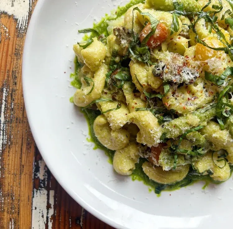 Overhead: Green pesto pasta with cheese, herbs, and tomatoes on wood.