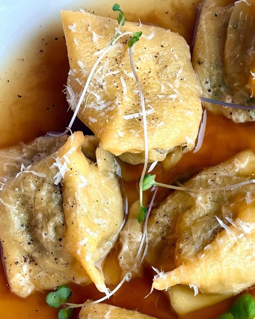 Tortellini-like pasta in dark sauce, topped with cheese, pepper, microgreens.