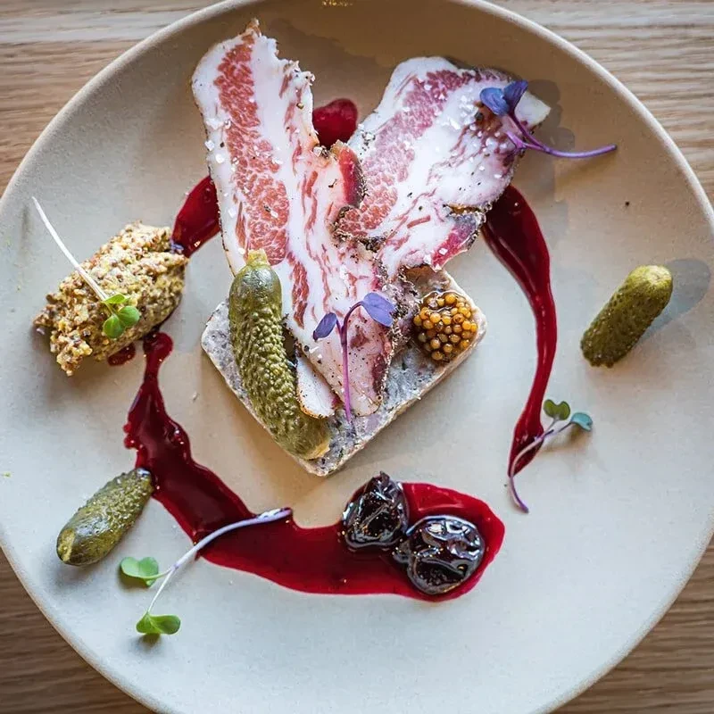 Elegant gourmet appetizer: lardo, gherkins, mustard, and dark red sauce.
