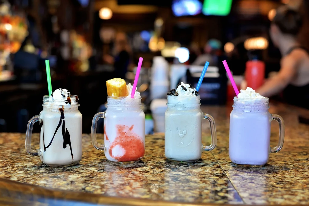 Four colorful, garnished milkshakes in mason jars on a bar.