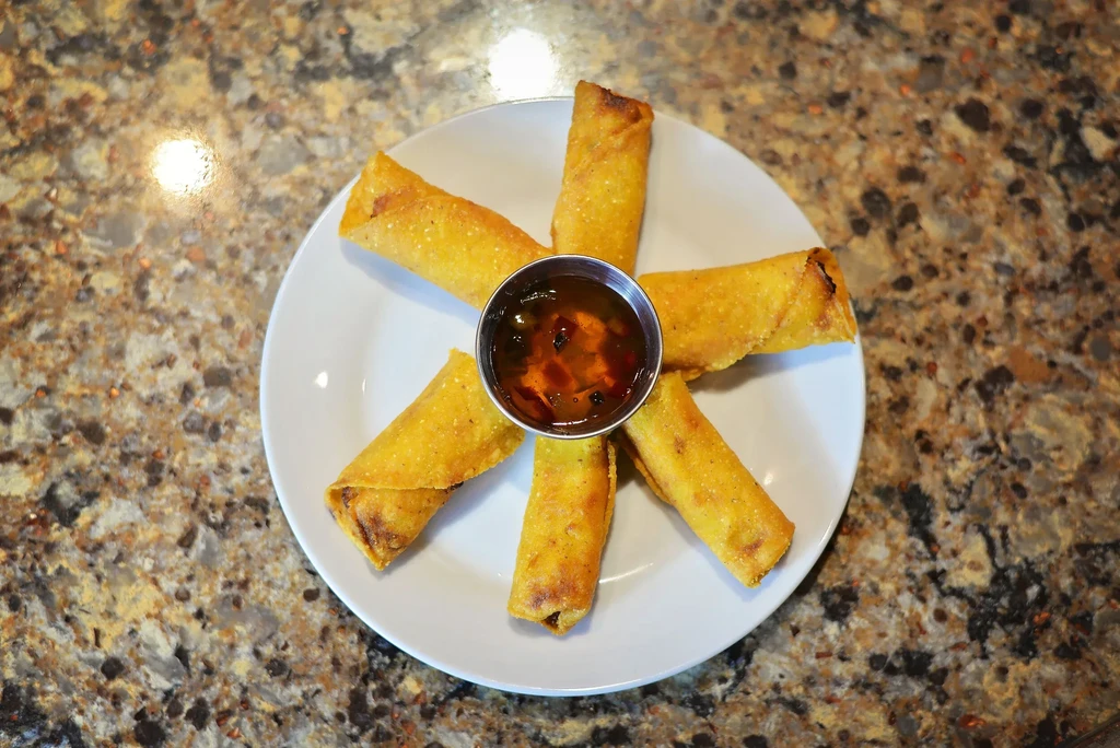 Six golden-brown egg rolls arranged around chili sauce on a plate.