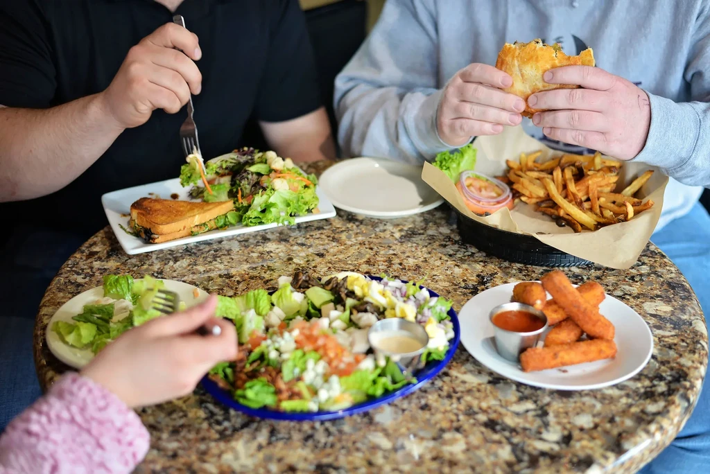 Hands eating salad, sandwiches, fries, and mozzarella sticks at a table.