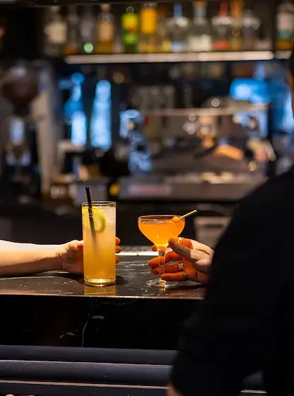 Two hands hold two garnished cocktails at a dark bar.
