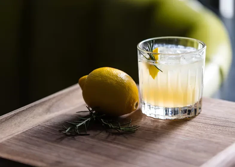 Cocktail with lemon, rosemary, and ice next to a lemon on wood.