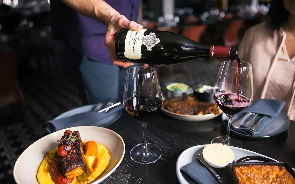 A hand pours Terra Grecale red wine among fine dining dishes.