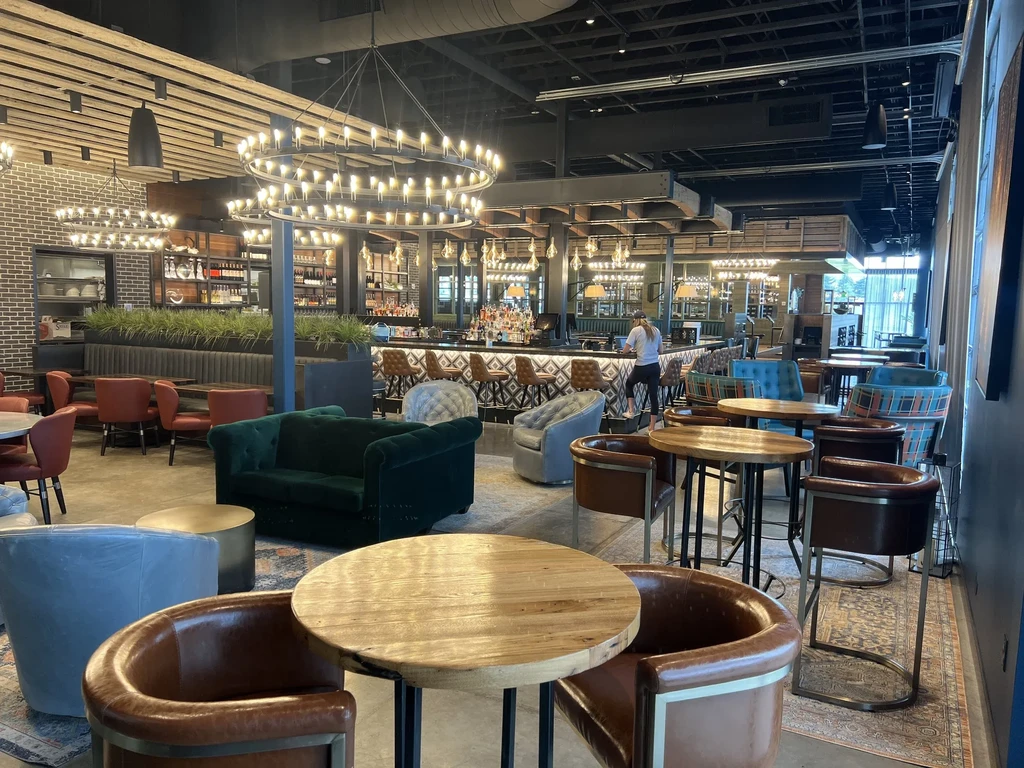 Modern restaurant bar with varied seating, large chandeliers, and exposed brick.