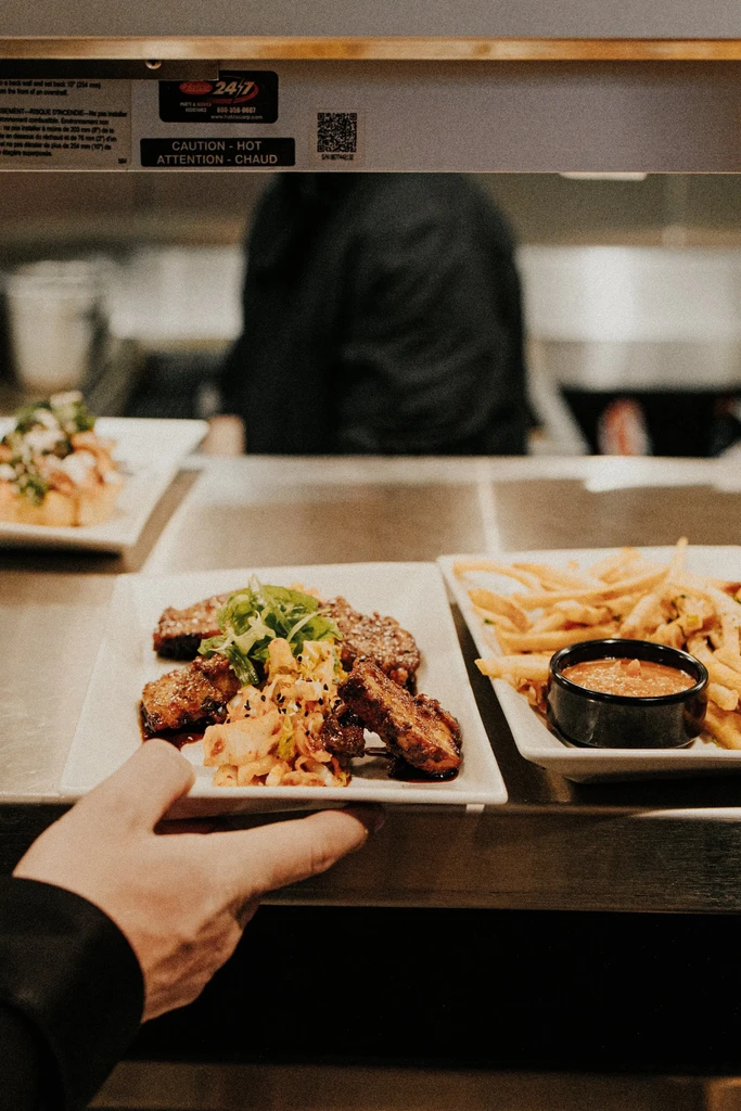 Hand holding glazed meat, kimchi, and fries at a kitchen pass.