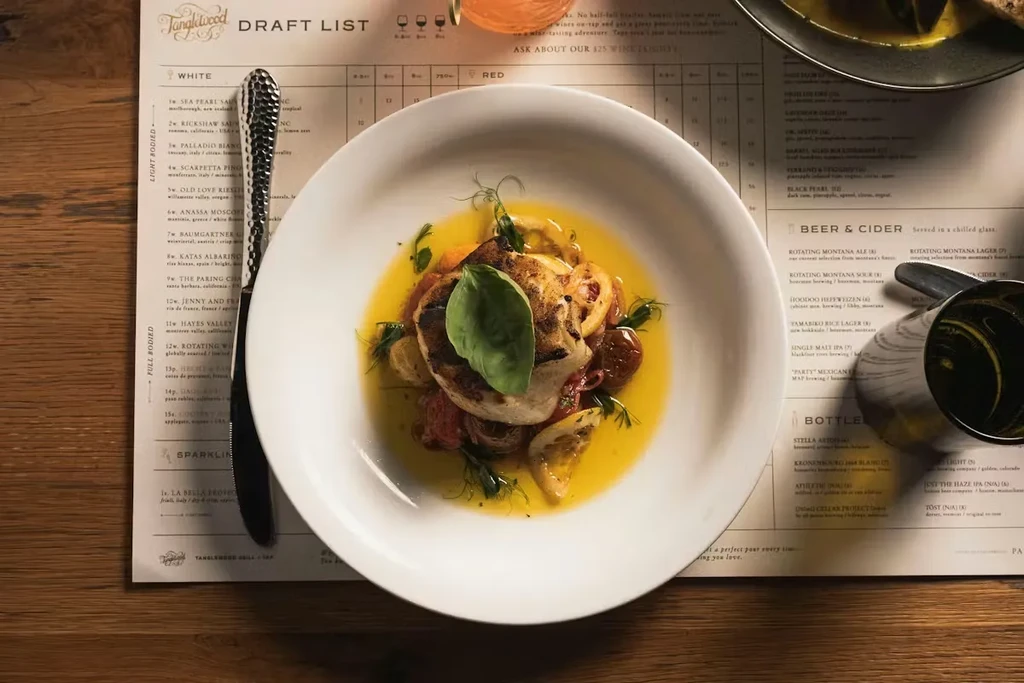 Overhead: Plated fish, heirloom tomatoes, fork, and Tanglewood menu on wood.
