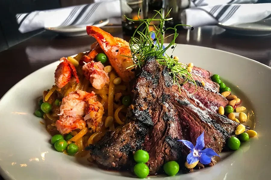 Grilled steak, lobster, pasta, peas, and nuts on a white plate.