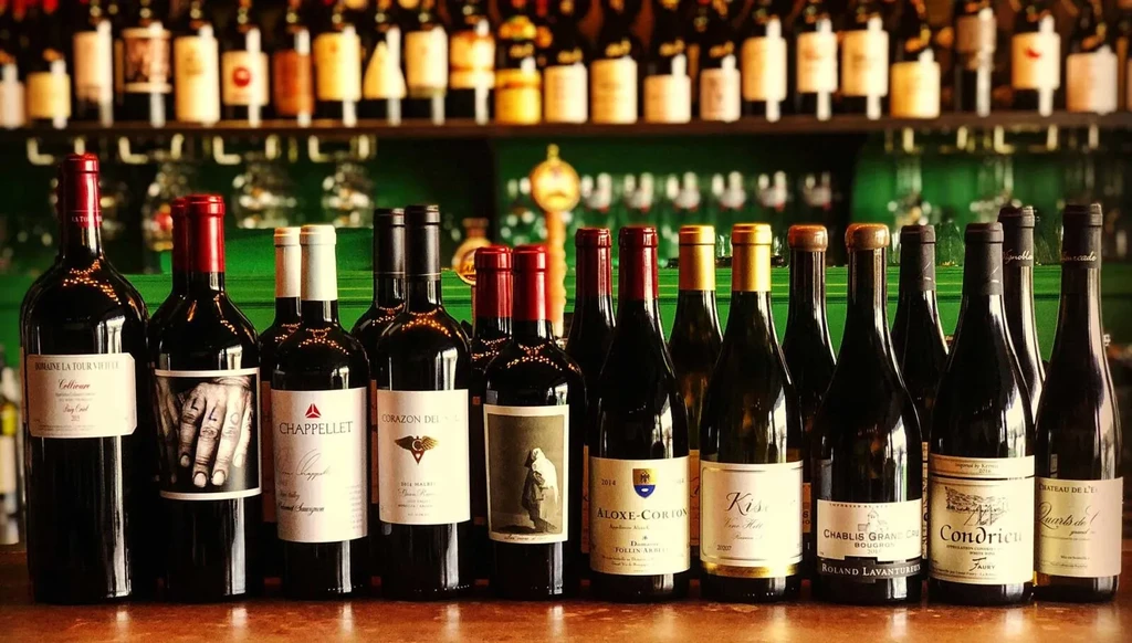 Numerous wine bottles on a wooden counter with a blurred shelf.