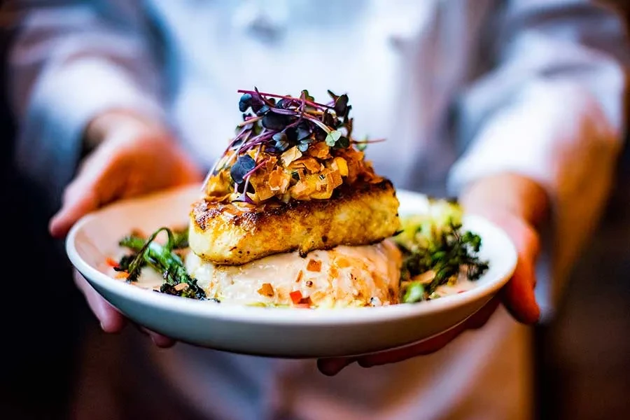 Chef holds plate of gourmet pan-seared fish with vegetables.
